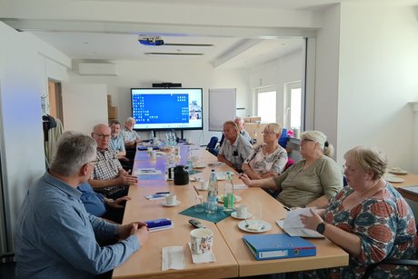 Mehrere Personen sitzen an einem langen Tisch vor einem Monitor und unterhalten sich.
