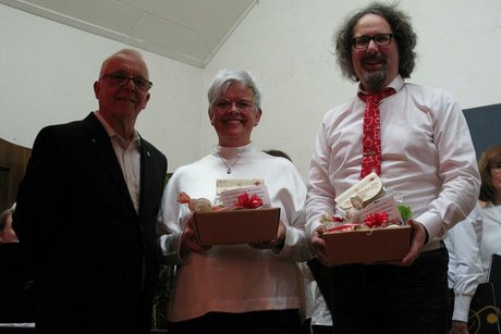 Drei Menschen stehen nebeneinander. Zwei davon halten Präsentkörbe in der Hand.