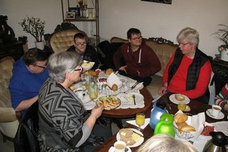 Fünf Menschen sitzen auf Sesseln und Sofa um einen Tisch herum und frühstücken.