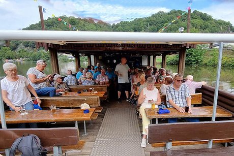 Viele Menschen sitzen gemeinsam auf Bänken an rustikalen Holztischen auf dem Floß unter einem Sonnendach.