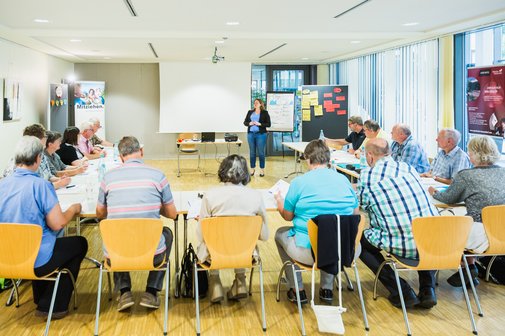 Menschen sitzen in einem Seminarraum an u-f&ouml;rmig aufgestellten Tischen. Vor Kopf steht eine Referentin vor einem White-Board.