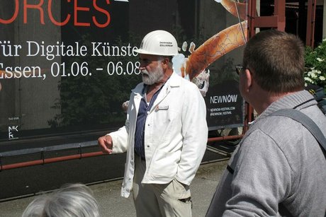 Ein bärtiger Mann mit weißem Bauhelm und weißer Bergmannsjacke steht vor der Reisegruppe und erzählt etwas. Im Hintergrund wirbt ein Plakat für eine Kunstausstellung zum Thema "Digitale Künste".