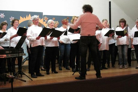 Der Chorleiter dirigiert singende Frauen, die auf einer Bühne stehen.