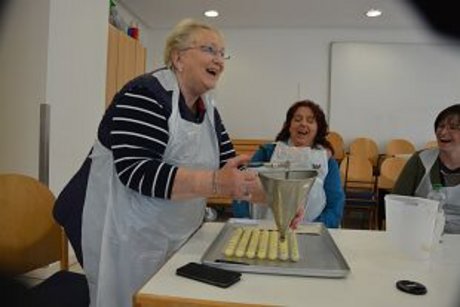 Eine Frau befüllt lachend weiße Schokoladenkugeln. Zwei weitere Frauen am Tisch lachen ebenfalls.