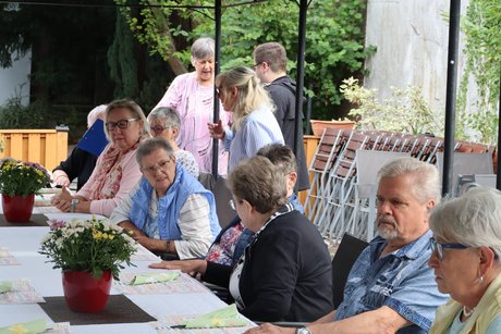 Grillnachmittag vom VdK Ortsverband Altendorf gemeinsam mit dem Ortsverband Borbeck. Teilnehmer sitzen an einem langen Tisch und unterhalten sich. Die Vorsitzende Andrea Grimm steht hinter einer Teilnehmerin und begrüßt sie.