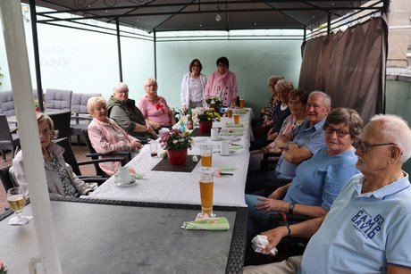 Grillnachmittag vom VdK Ortsverband Altendorf gemeinsam mit dem Ortsverband Borbeck. Teilnehmer des Ortsverband Borbeck sitzen an einem langen Tisch. 2 Frauen stehen am Ende des Tisches und begrüßen die Teilnehmer.