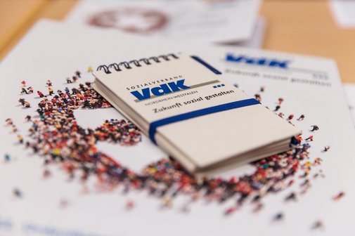Ein Notizblöckchen mit blauem VdK-Logo liegt auf einer buntgemusterten Broschüre.