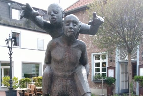 Die Skulptur in Viersen-Dülken zeigt einen blinden Mann, der einen Mann auf dem Rücken trägt. Der Mann auf dem Rücken zeigt nach vorne und sagt dem Blinden den Weg.