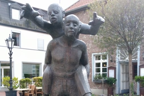 Die Skulptur in Viersen-Dülken zeigt einen blinden Mann, der einen Mann auf dem Rücken trägt. Der Mann auf dem Rücken zeigt nach vorne und sagt dem Blinden den Weg.