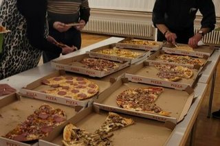 Acht Kartons mit verschiedenen Pizzen stehen auf einem Tisch. Die Gäste, hier im Bild zwei Männer und eine Frau, bedienen sich selbst.