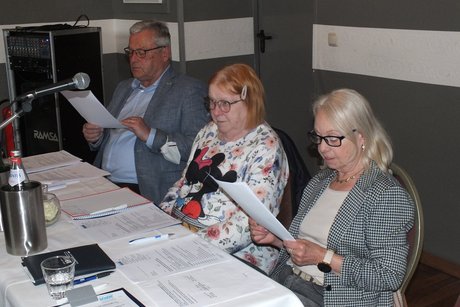 Jahreshauptversammlung 2026 Ein Mann und zwei Frauen sitzen vor Dokumenten und lesen von Textblättern vor.