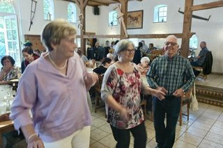 Drei Menschen tanzen gemeinsam. Im Hintergrund sitzen Menschen an langen Tischen.