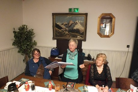 Zwei Frauen sitzen und die dritte Frau in der Mitte steht an einem gedeckten Tisch und spricht.