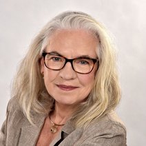 Auf dem Foto ist Frau Busch abgebildet. Frau Busch hat weißblonde, schulterlange Haare, trägt eine Brille und einen beigen Blazer