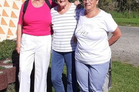Drei Frauen stehen Arm in Arm vor der Waffelherz-Skulptur auf einem Stück grünem Rasen.