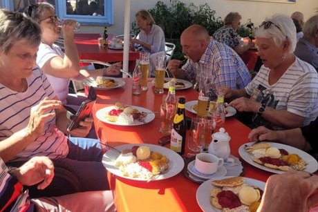 Eine Gruppe Menschen sitzt gesellig versammelt unter einem großen Sonnenschirm an einem langen Tisch auf der Außenterrasse des Waffelhauses und lässt sich Waffeln und Kaltgetränke schmecken.