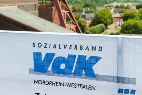 VdK-NRW-Banner an einem Zaun, dahinter der F&ouml;rderturm der Zeche Zollverein und viele Baumwipfel.