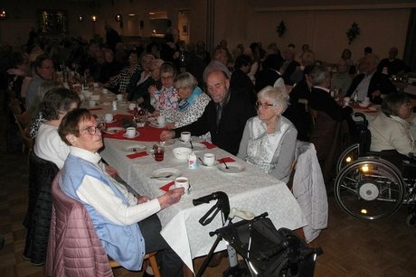 Viele Menschen sitzen in einem Saal an langen, weihnachtlich geschmückten Tischen.