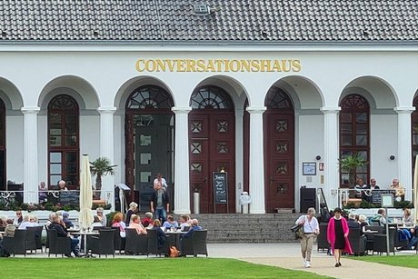 Außenfassade eines Gebäudes mit großem Säulengang und Bogenfenstern. In goldenen Buchstaben steht Conversationshaus über den Bögen. Vor dem Gebäude sitzen viele Menschen in schwarzen Korbstühlen an Tischen.