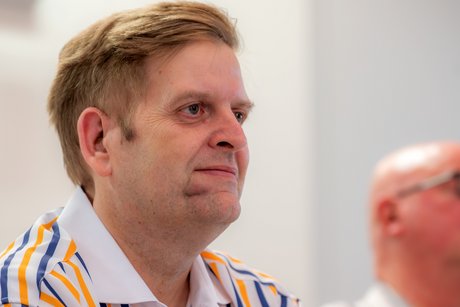 Ein Mann mit blonden kurzen Haaren und gestreiftem Hemd schaut rechts aus dem Bild heraus.