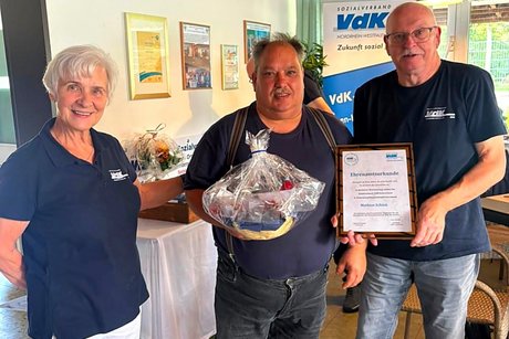 Jubilars- und Ehrenamts-Verleihung des VdK-Ortsverbandes Essen - Katernberg - Stoppenberg - Schonnebeck. Eine Frau und ein Mann übergeben an einen anderen Mann eine urkunde, eine Ehrennadel und ein Präsent.