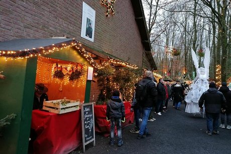 Heimelig beleuchtete Weihnachtsmarktbuden reihen sich entlang der Ziegelfassade eines Hauses auf. Besucher schlendern hier unter Bäumen die Buden entlang