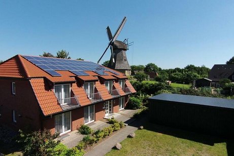 Links im Bild ein rotverklinkertes Gebäude mit Solarpaneelen auf dem Dach, im Hintergrund eine alte Windmühle.