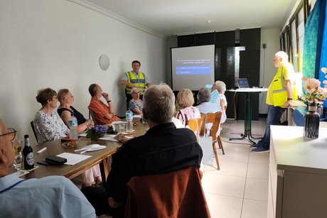 Verkehrsquiz mit Polizeihauptkommissar Hans-Joachim Ruhl. Mehrere Personen sitzen an einem langen Tisch und hören Polizeihauptkommissar Hans-Joachim Ruhl zu.