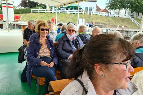 Menschen sitzen gemeinsam auf Holzbänken an Deck eines Ausflugsschiffs unter einer Markise.