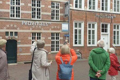 Menschen stehen gemeinsam auf einem Platz vor rotverklinkerten Gebäuden mit der Aufschrift Pelzerhaus.