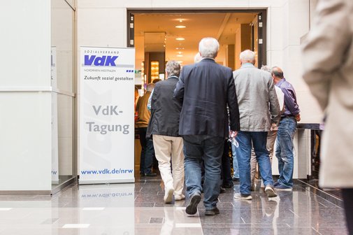 Menschen stehen im Eingangsbereich eines Geb&auml;udes in einer Schlange. Links von ihnen ein Banner mit VdK-NRW-Logo und der Aufschrift VdK-Tagung