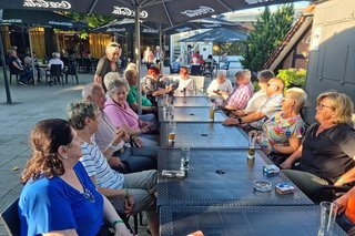 Eine große Gruppe Menschen sitzt gemeinsam bei Getränken an einer langen Tischreihe unter Sonnenschirmen auf der Außenterrasse des Ferienressorts.