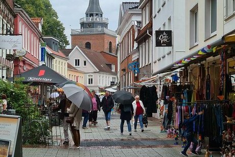 Menschen mit Regenschirmen laufen durch die Fußgängerzone von Aurich. Im Hintergrund ragt der Turm der Lambertikirche in den wolkenverhangenen Himmel.