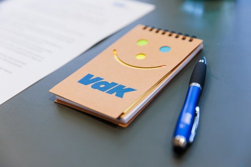 Ein brauner VdK-Block mit Logo und Lachgesicht und ein blauer VdK-Kugelschreiber liegen auf einem Tisch.
