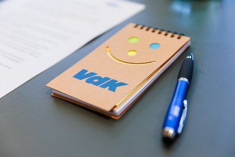Ein brauner VdK-Block mit Logo und Lachgesicht und ein blauer VdK-Kugelschreiber liegen auf einem Tisch.