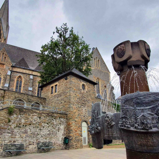 Historische Steinkirche mit spitzem Turm, umgeben von einer Mauer und einem Brunnen mit Reliefs und Wasserspiel.