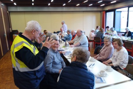 Viele Menschen sitzen an langen Tischen. Ein Mann mit gelber Warnweste spricht zu den Menschen.