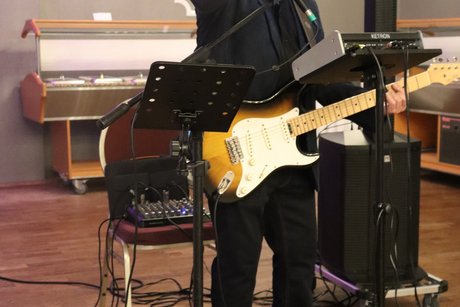 Ein Mann mit Gitarre steht vor einem Mikrophone und zeigt mit der Hand zur Kamera.