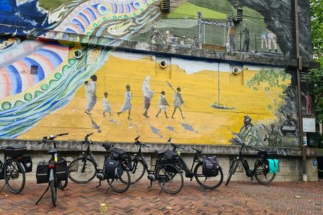 Fahrräder parken entlang einer hohen Mauer, auf der ein beeindruckendes Mureal gesprayt ist, das einen bunten Fluss und Menschen in der Wüste zeigt.
