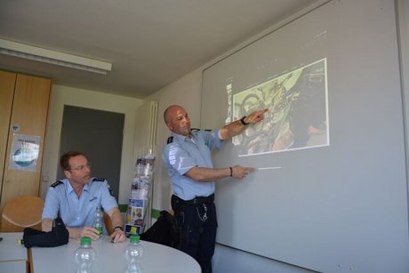 Ein Polizist sitzt am Tisch, ein anderer steht vor einer PowerPoint-Präsentation und erklärt, was dort zu sehen ist.