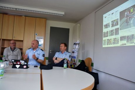 Zwei Polizisten sitzen an einem Tisch, neben ihnen eine PowerPoint-Präsentation an der Wand