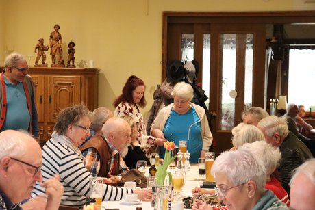 Einige Menschen sitzen an einem langen Tisch und andere stehen am Tisch und reden miteinander.
