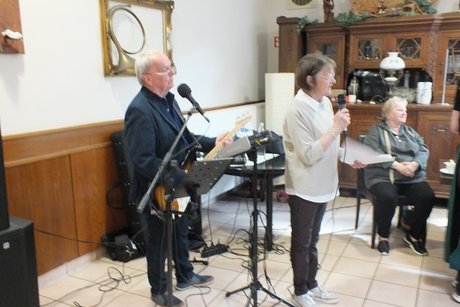 Eine Frau spricht in ein Mikrophone und hinter Ihr steht ein Musiker mit Gitarre.