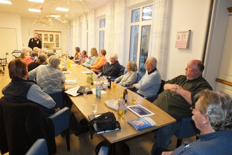 Ein Mann steht vor einem langen Tisch, an dem viele Menschen sitzen und ihm zuh&ouml;ren.