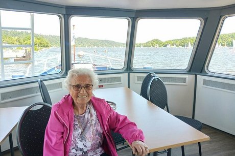 Eine alte weißhaarige Frau in rosa Bluse und pinker Jacke sitzt an einem Tisch an Deck eines Schiffes. Durch die Fenster im Hintergrund blickt man auf den Baldeneysee und das bewaldete Ufer.