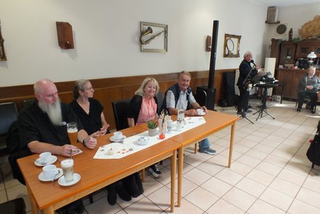 4 Personen sitzen an einem Tisch und im Hintergrund ist ein Musiker zu sehen.