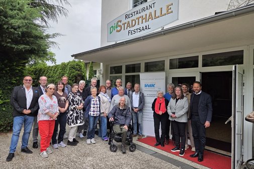 Eine große Gruppe Menschen steht vor dem Eingang zu einem Lokal rechts und links von einem Roll-Up mit VdK-Logo. Darüber ein Schild Restaurant - Die Stadthalle. Festsaal.