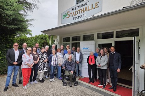 Eine große Gruppe Menschen steht vor dem Eingang zu einem Lokal rechts und links von einem Roll-Up mit VdK-Logo. Darüber ein Schild Restaurant - Die Stadthalle. Festsaal.