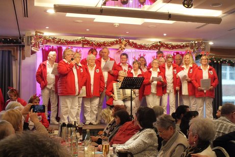 Ein Chor steht auf einer B&uuml;hne in wei&szlig;er Kleidung mit roten Jacken und singen.