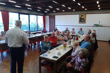 Mehrere Menschen sitzen an langen Tischen und h&ouml;ren einem Beamten in Uniform zu.